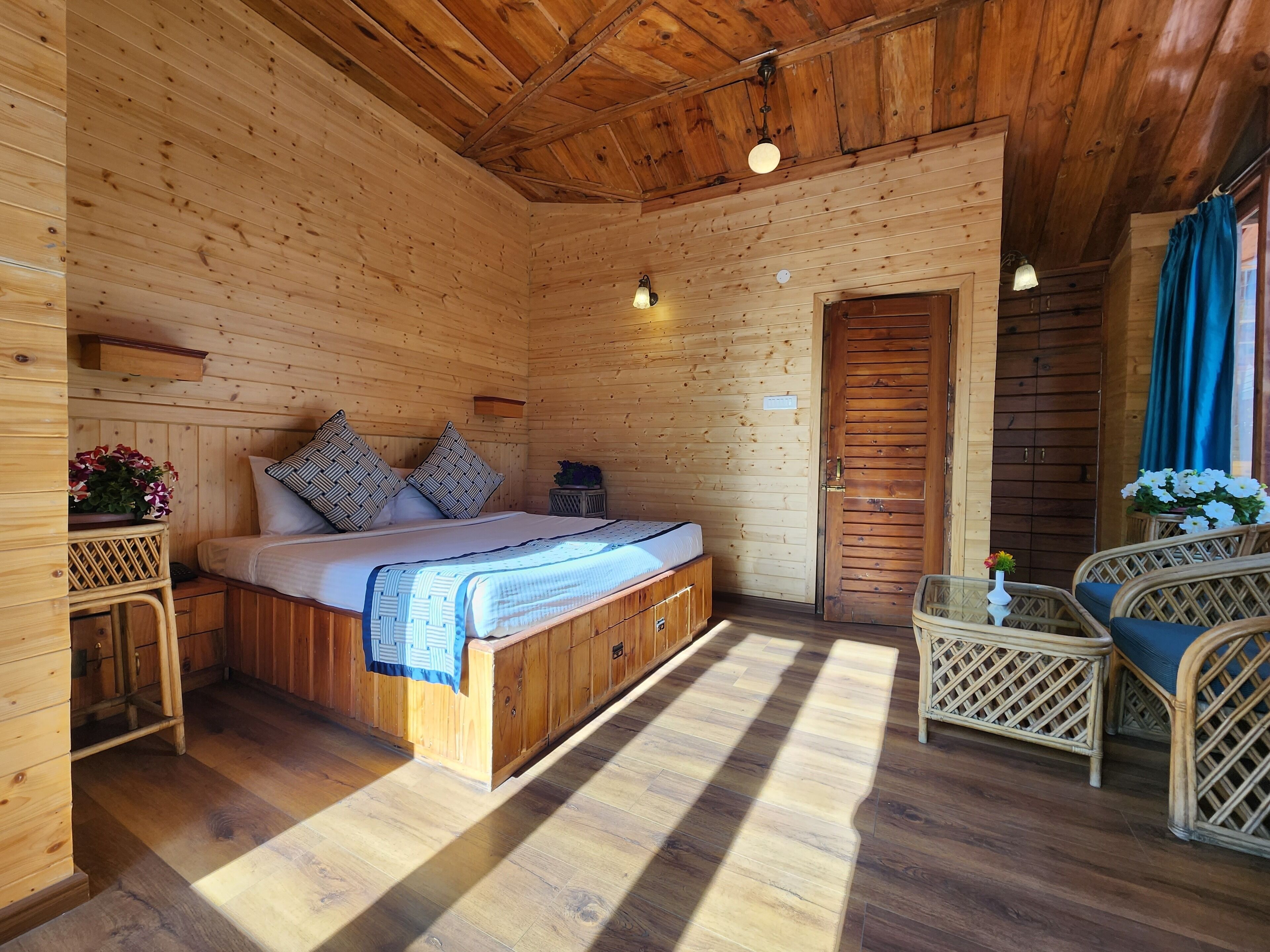 A wooden cabin room with a bed, a small table, wicker furniture, and a door leading outside.