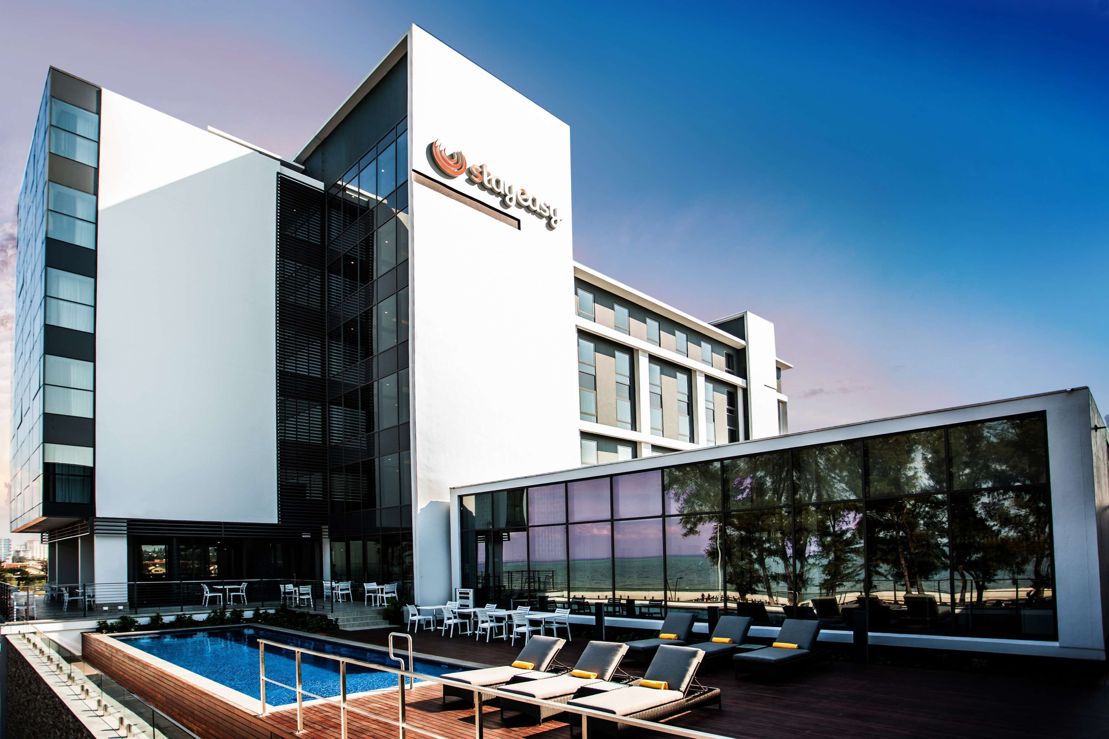 A modern hotel with a rooftop pool area, outdoor seating, and large windows offering views of the sea.