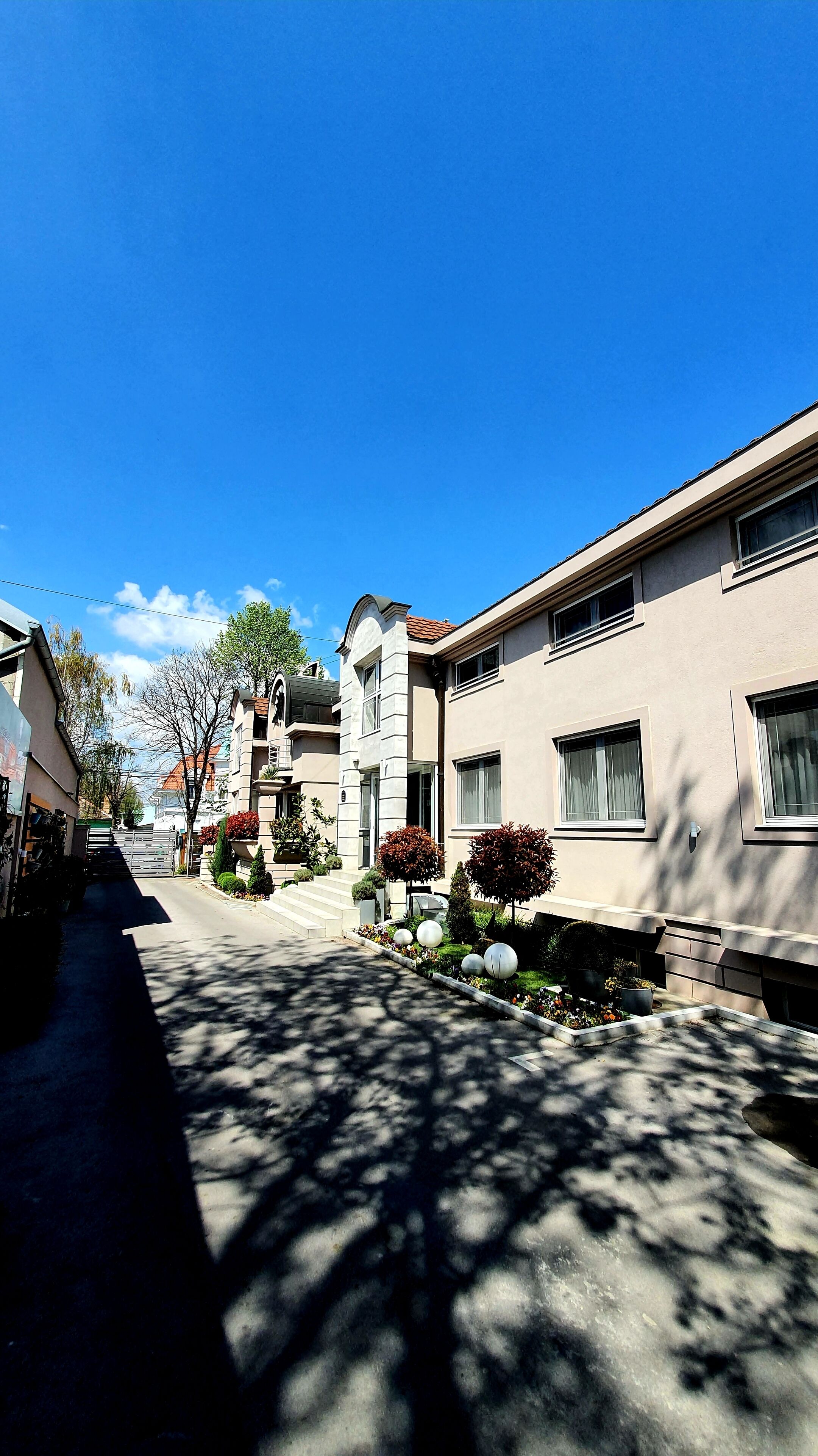 Eine Wohnstraße mit modernen Häusern, wolkenlosem Himmel und Baumschatten auf dem Bürgersteig.