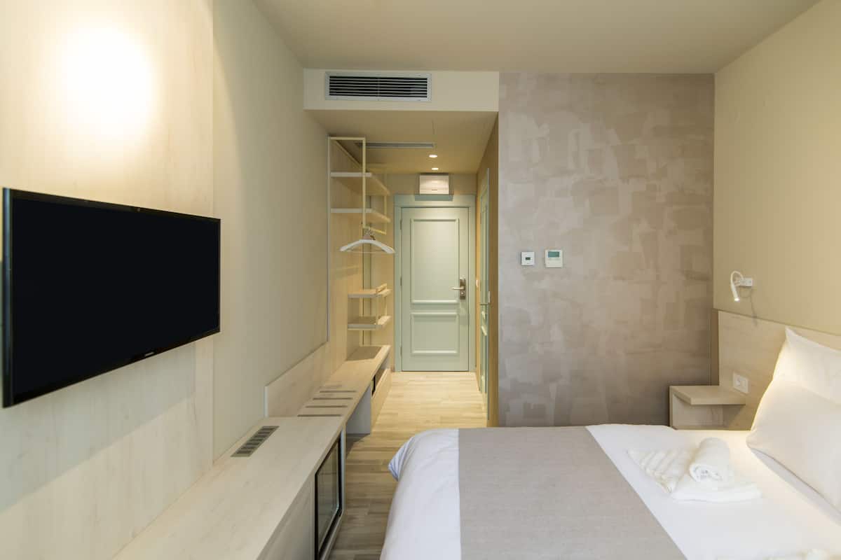 A modern hotel room with a flat-screen TV mounted on the wall, a bed with a neatly made white comforter, a bedside table, and a small bedside lamp.