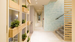Un interior moderno con un largo pasillo, estanterías de madera con plantas, una escalera con barandillas de madera y luces empotradas en el techo.