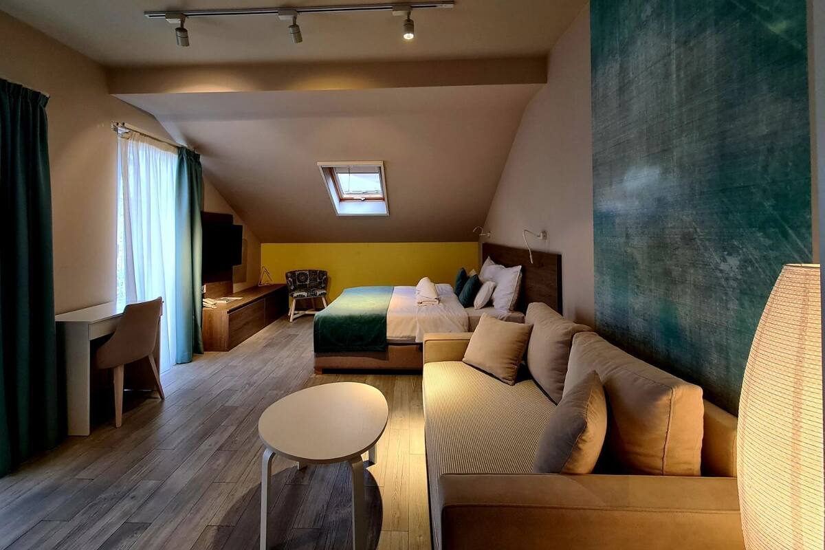 A modern hotel room with a loft bed, a sofa, a small table, and a desk. There is a skylight above the bed and a large abstract painting on the wall.