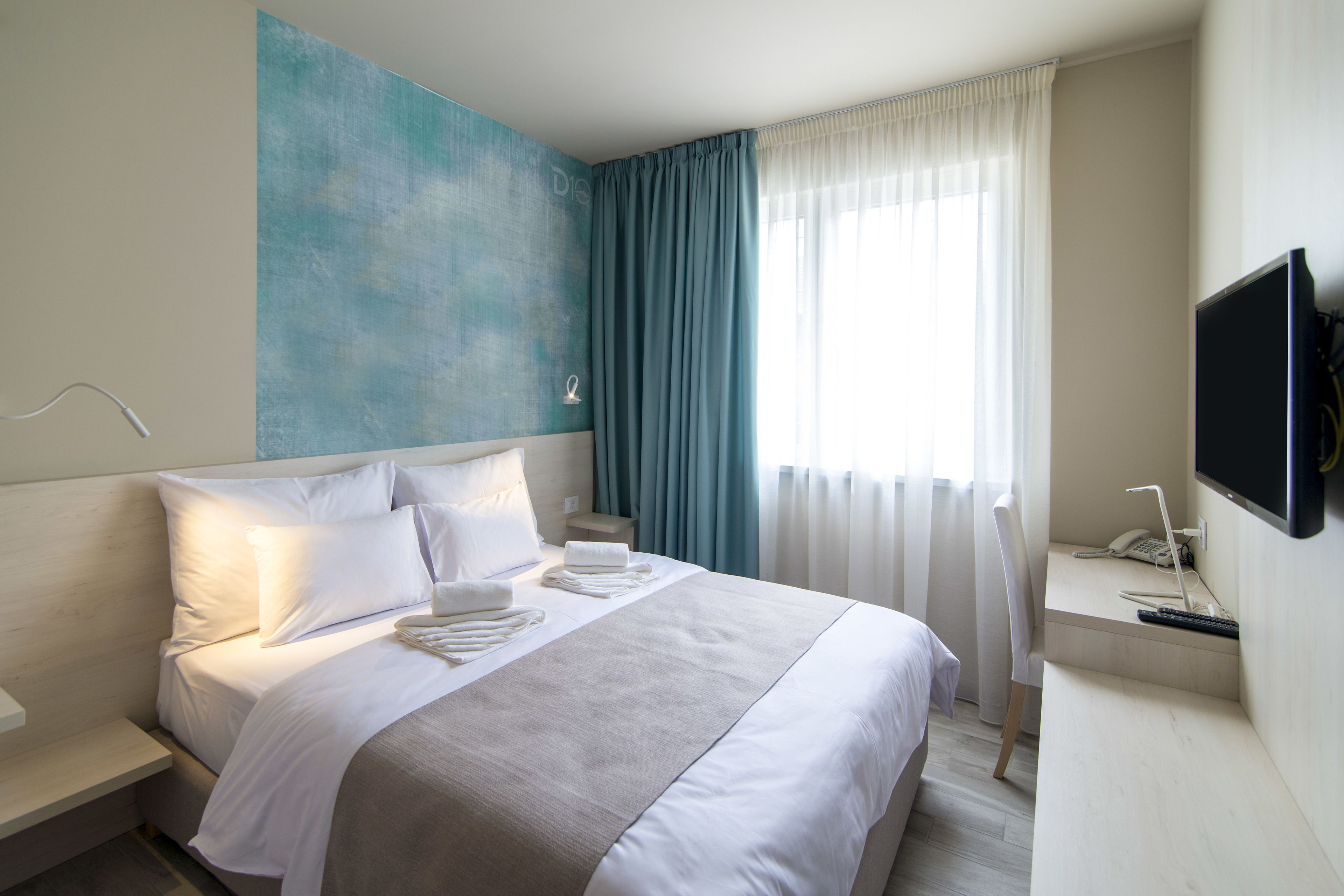 Une chambre d’hôtel comprenant un lit, un téléviseur à écran plat, une table de chevet, une petite table, une lampe fixée au mur et une fenêtre avec des rideaux.
