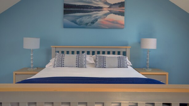 Een slaapkamer met een bed, twee bedlampjes, een blauwe wand en een schilderij van een berg en meer.