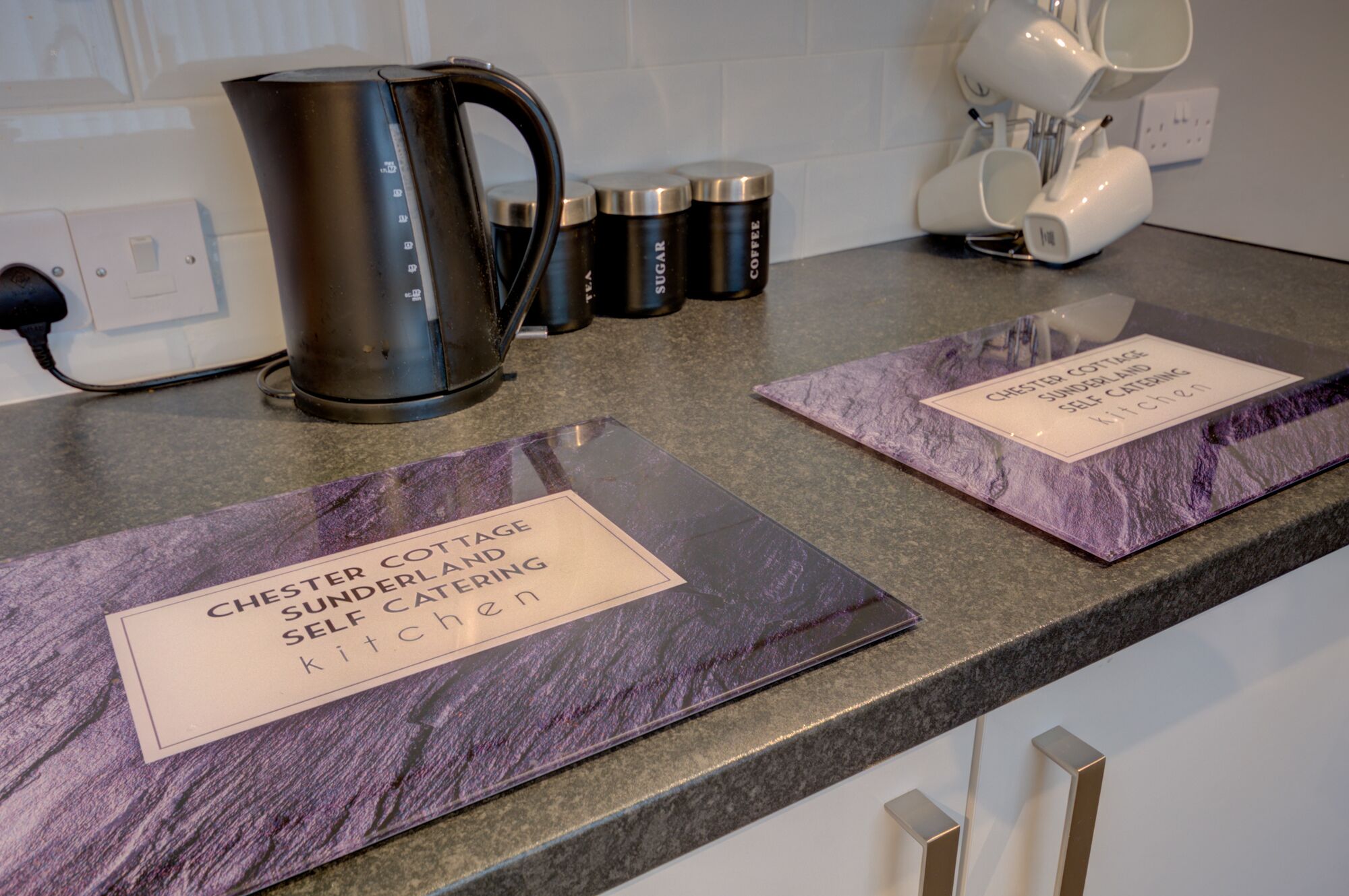 A kitchen counter with a kettle, coffee mugs, and self-catering signs.