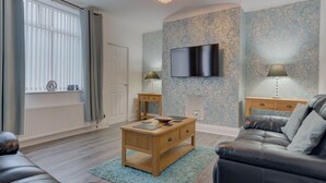 Een woonkamer met een houten salontafel, een leren bank, een aan de muur gemonteerde televisie en behang met een patroon.