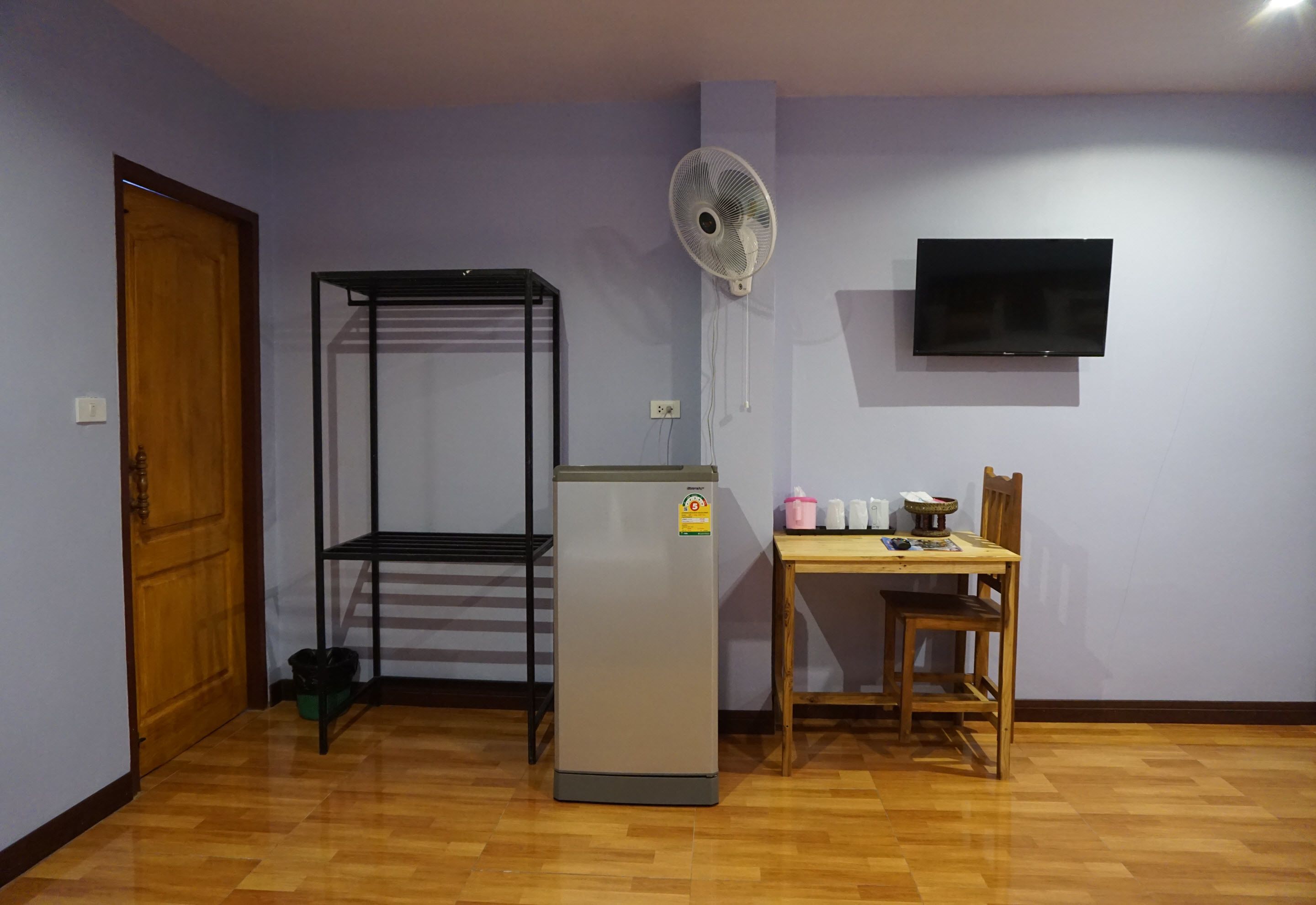 Una habitación con puerta de madera, estantería metálica, refrigerador, mesa con silla, ventilador de pared y televisor.