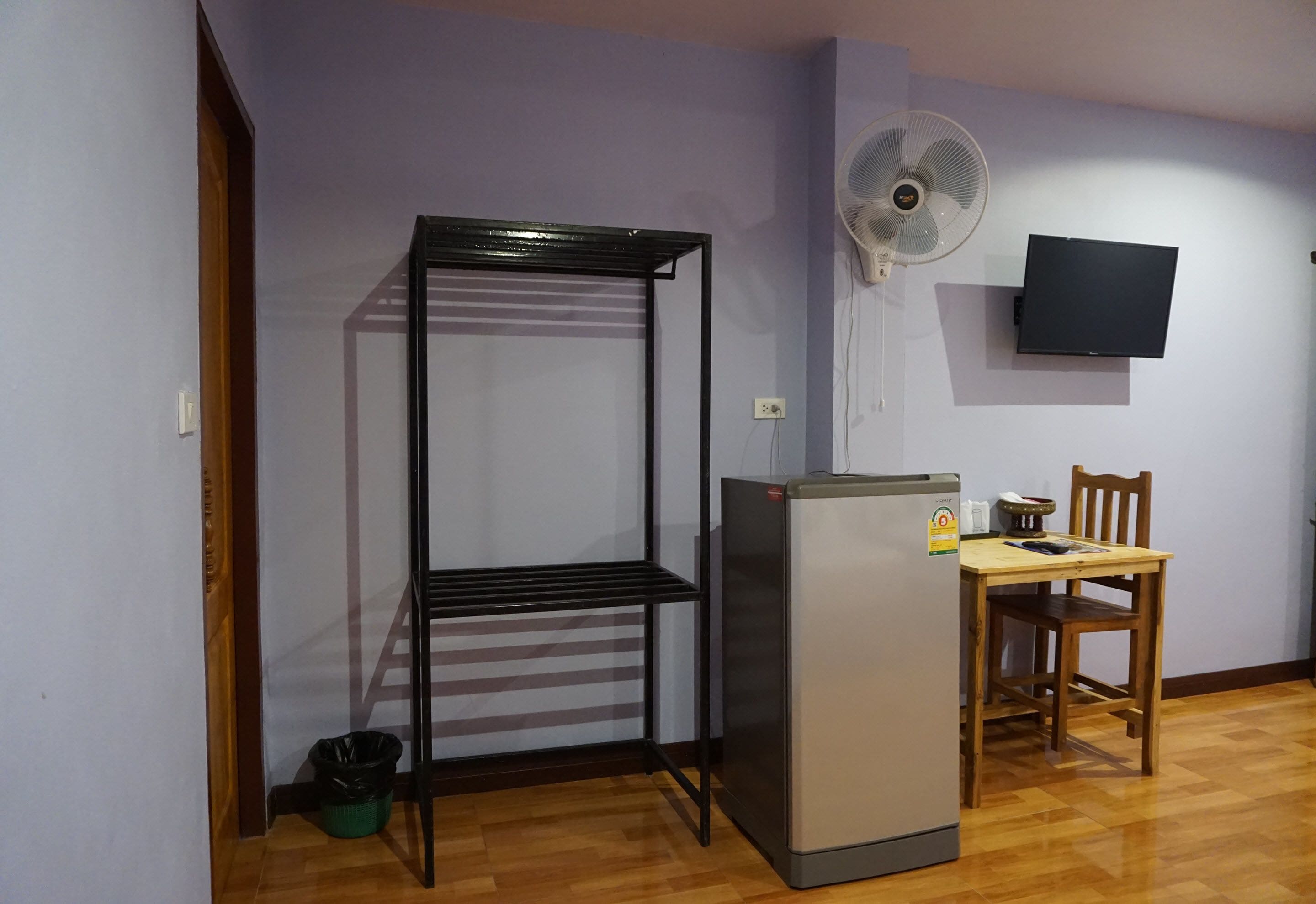 Una habitación con una estantería metálica, un refrigerador, una mesa de madera con una silla, un ventilador de pared y un televisor.
