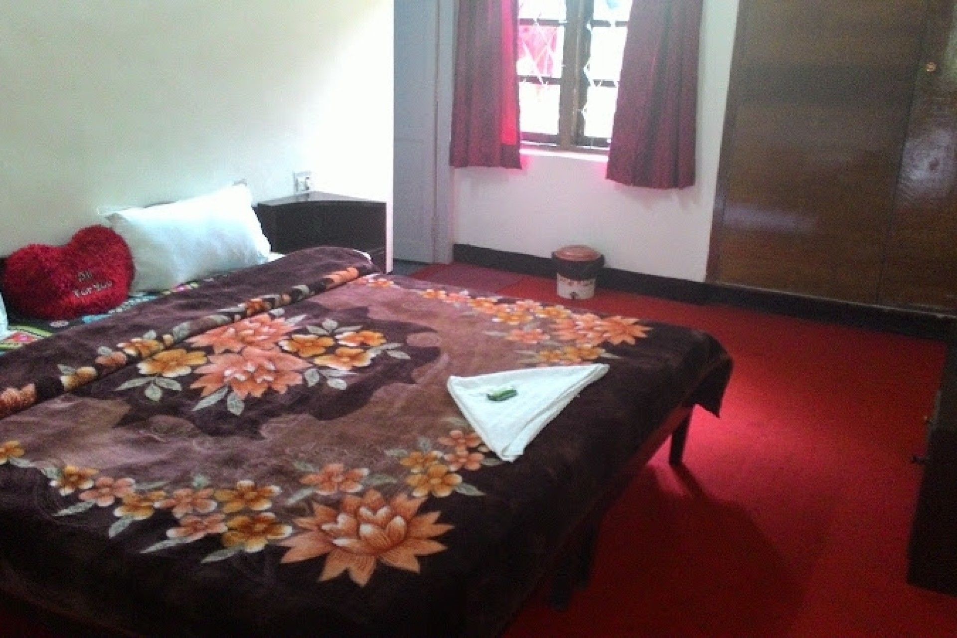 A bedroom with a floral bedspread, a red heart-shaped pillow, a small bedside table with a lamp, a window with red curtains, and a red carpeted floor.