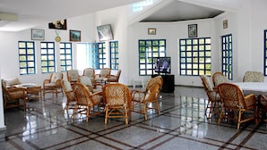 A spacious room with a high ceiling, large windows with blue frames, wicker furniture, and a tiled floor.