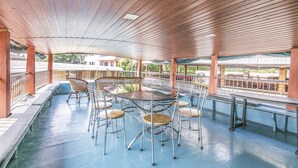 An outdoor covered patio with metal chairs and tables, a blue floor, and a view of buildings in the background. - GuestHouser 3 BHK Houseboat 147b (Ambalapuzha)
