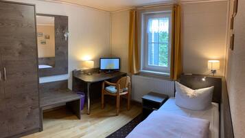 A compact hotel room with a bed, a desk with a computer, a chair, a small table, and a window with curtains.