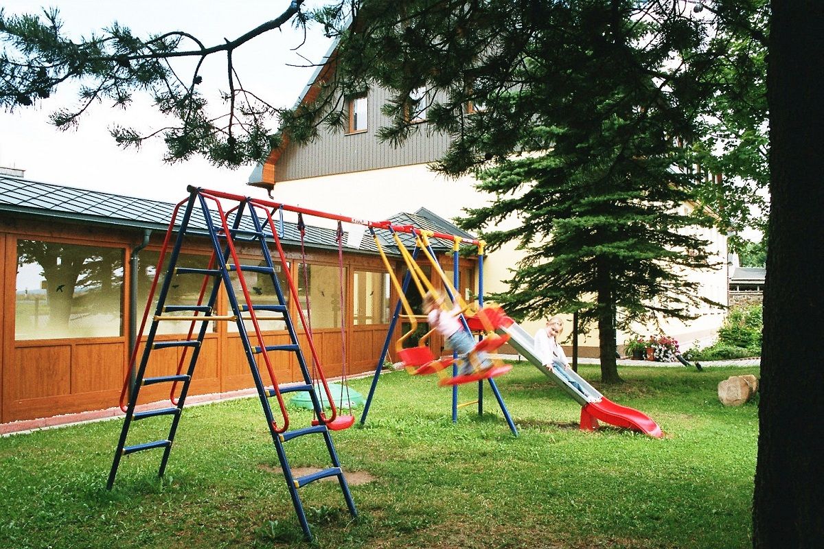 Un parque infantil con tobogán y columpios, rodeado por una valla de madera y una casa al fondo.