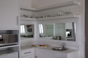 Cocina compacta con muebles blancos, microondas integrado, lavamanos y un espejo sobre la encimera.