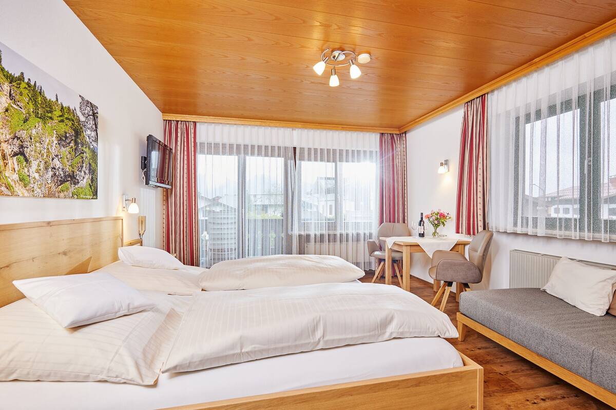 Une chambre d’hôtel avec un plafond en bois, un lit avec une literie blanche, un canapé, un bureau avec une chaise et une grande fenêtre avec des rideaux.
