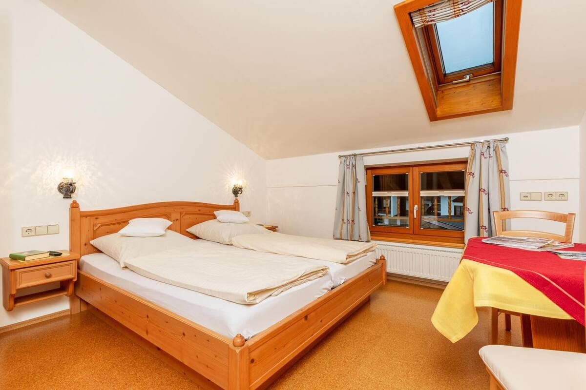 Une chambre à coucher avec un lit en bois, deux tables de chevet, une fenêtre avec des rideaux, une petite table recouverte d’une nappe rouge et une chaise.