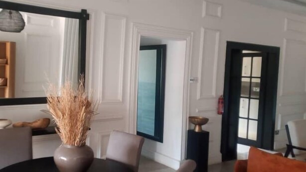 Een eethoek met een tafel, stoelen, een vaas met gedroogde planten, een lamp en een spiegel aan de muur.