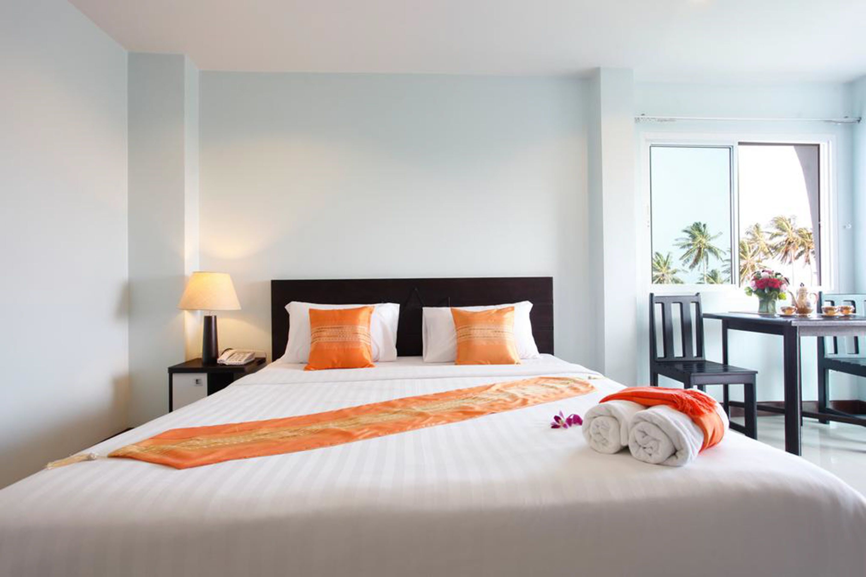 A hotel room with a bed, a nightstand with a lamp, a desk with chairs, and a view of palm trees through a window.