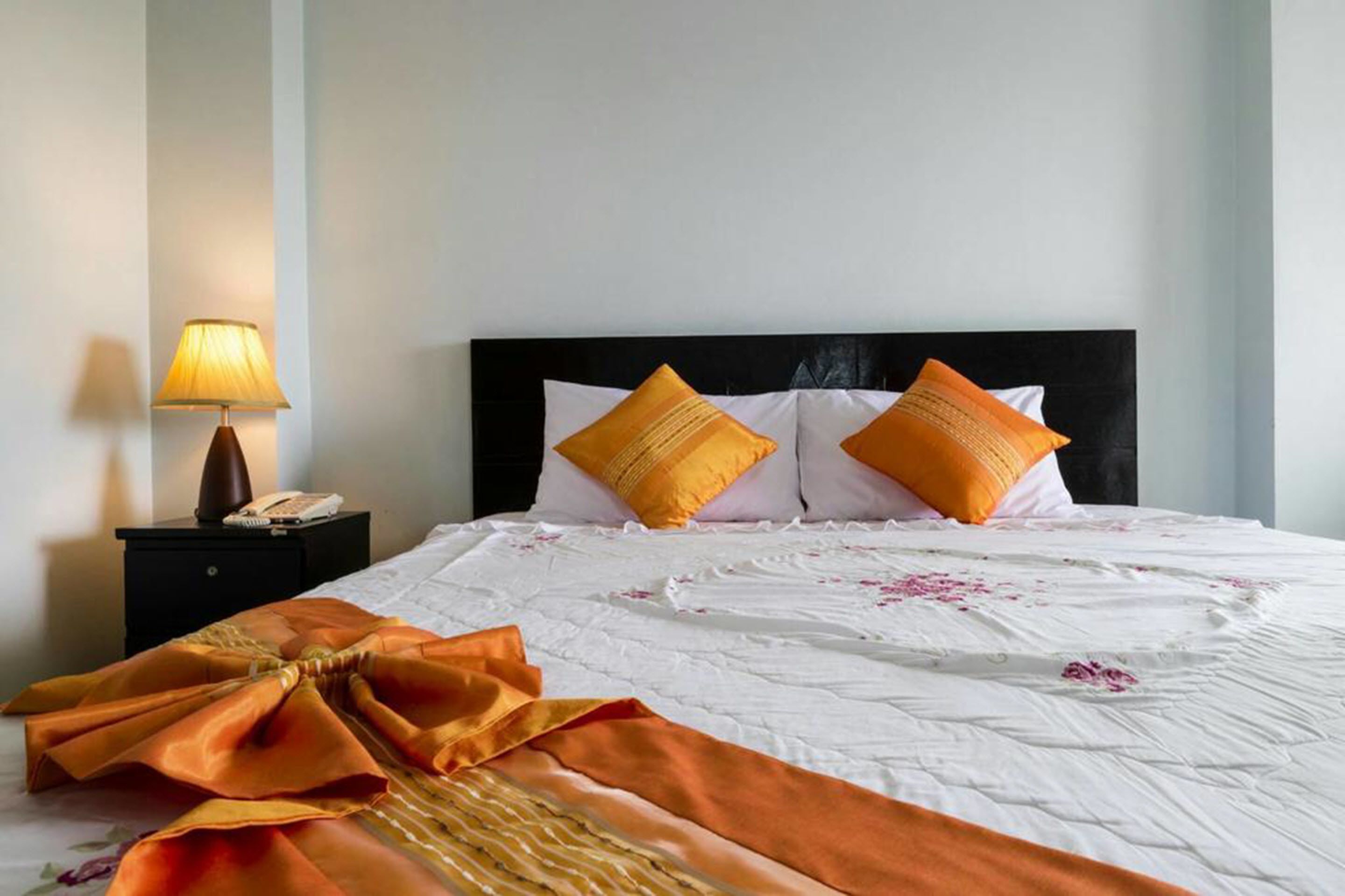 Een keurig opgemaakt bed met een witte sprei en twee oranje kussens, een bedlampje, en een opgevouwen oranje doek op het bed.