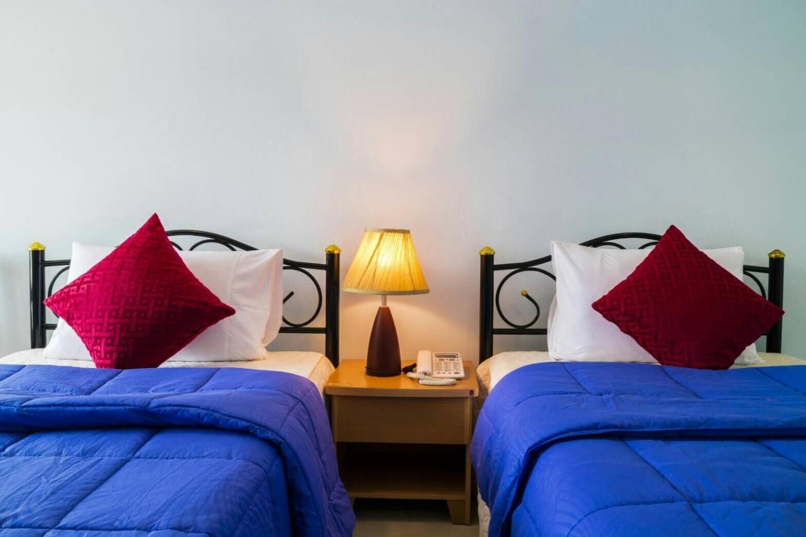 A hotel room with two beds, each with a blue bedspread and red pillows, a bedside table with a lamp and a telephone, and a plain wall in the background.