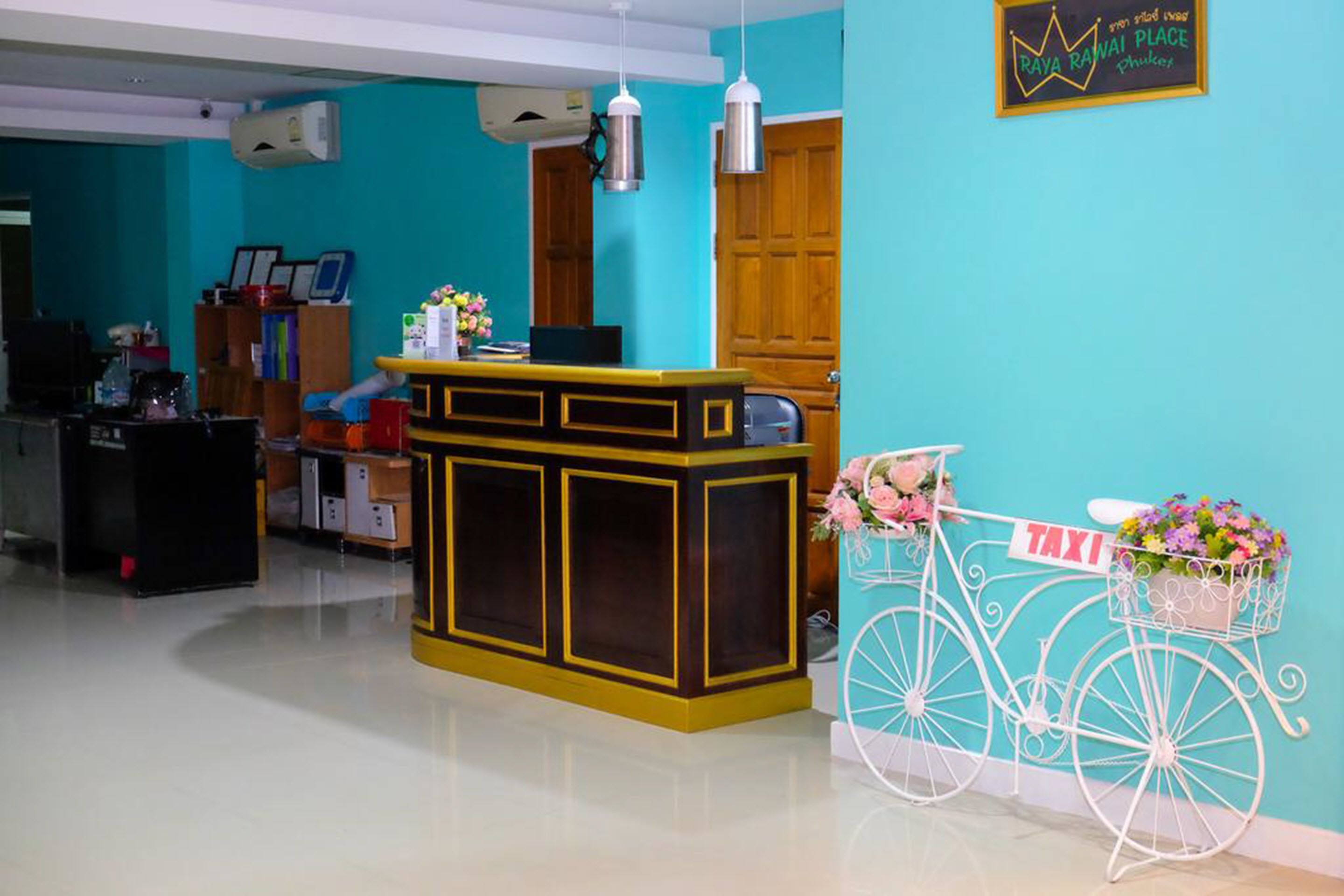 Un espace d’accueil avec un mur turquoise, un comptoir de réception, un vélo avec un panier rempli de fleurs et un panneau indiquant « TAXI ».