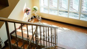 Una escalera con barandilla de metal que conduce a una plataforma. Hay un ventanal con persianas en la plataforma, y un jarrón con flores en el suelo.