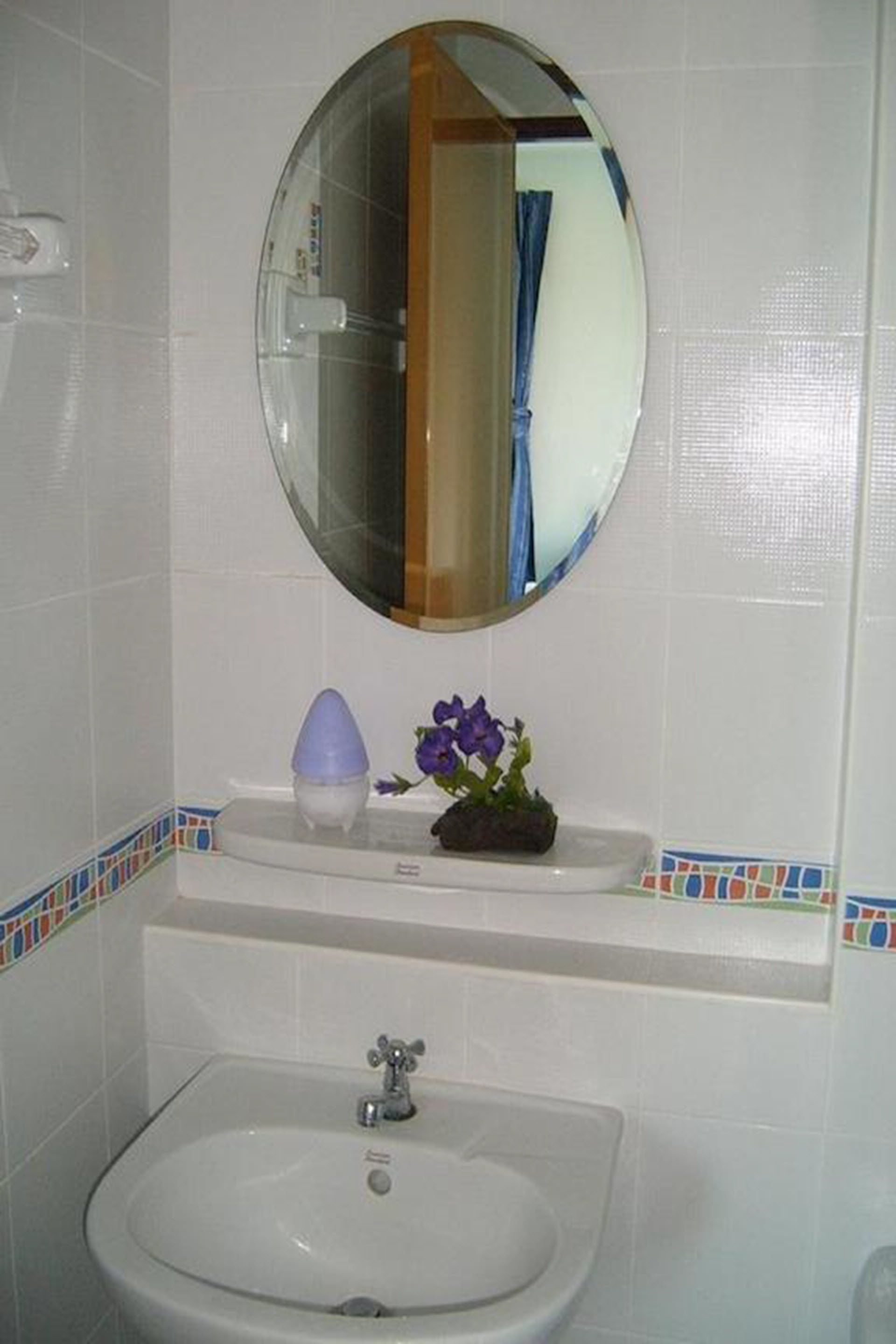 A bathroom with a white sink, a mirror above it, and a small shelf with a purple flower and a blue object.