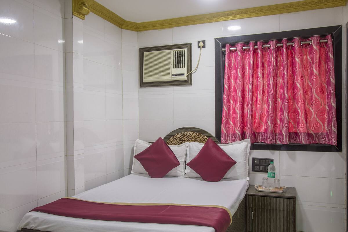 A hotel room with a bed, a nightstand, an air conditioning unit, a window with a curtain, and a wall-mounted light fixture.