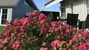 A building with a flagpole, a garden with pink flowers, and a clear blue sky. - Oldfruen De luxe (Vaeggerlose)