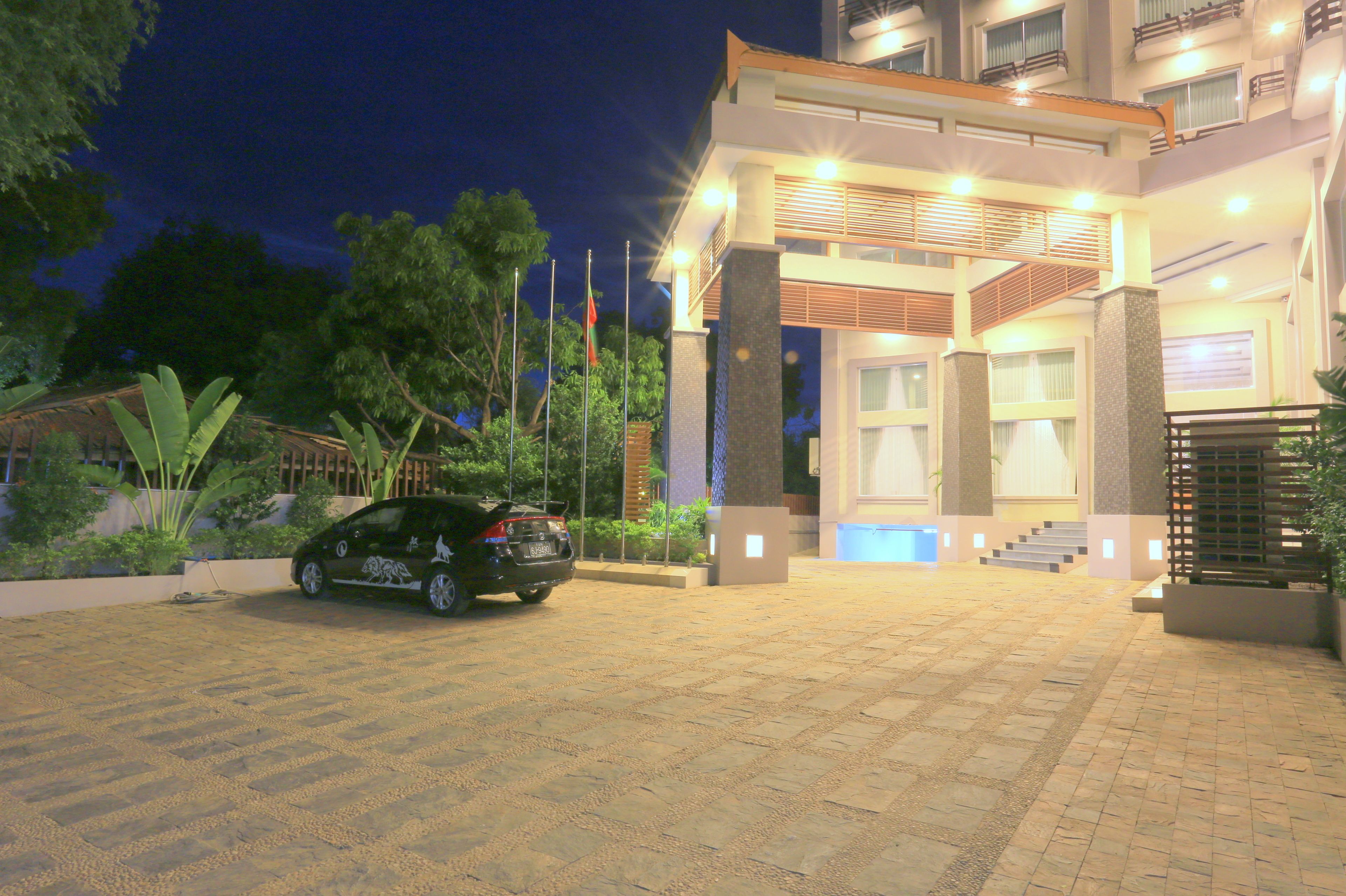 A hotel entrance with a paved driveway, a car parked in front, and a staircase leading to the entrance.