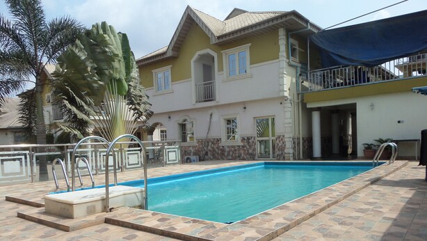 Una piscina con un borde de baldosas, una escalera metálica y una casa con balcón al fondo.