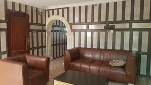 Una habitación con un sofá de cuero marrón, un sillón a juego y una mesita pequeña. Hay una puerta de madera y un amplio vano de arco.