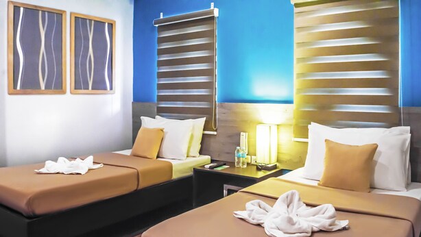 Habitación de hotel con dos camas, cada una con colcha marrón y almohadas blancas. Hay dos marcos de cuadros en la pared, una mesita de noche con lámpara y una ventana con persianas.
