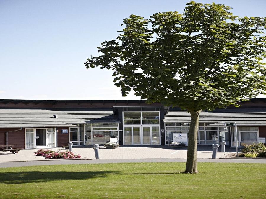 A modern building with a glass facade, a tree in the foreground, and a paved area with a bench.