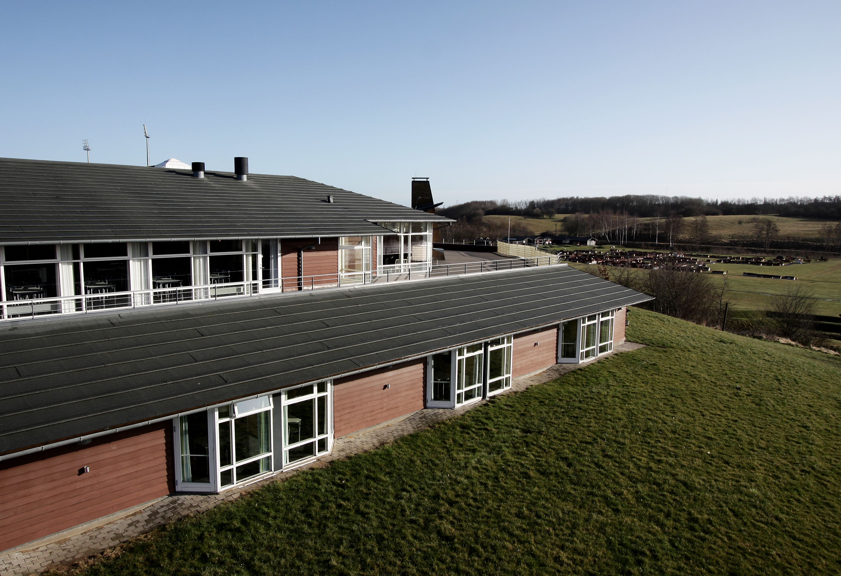 Een modern gebouw met een plat dak en grote ramen, omgeven door een gazon en een veld in de verte.