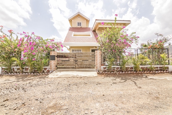 Una casa de dos pisos con techo de tejas rojas, un portón de entrada de ladrillo y plantas florecientes de color rosa.
