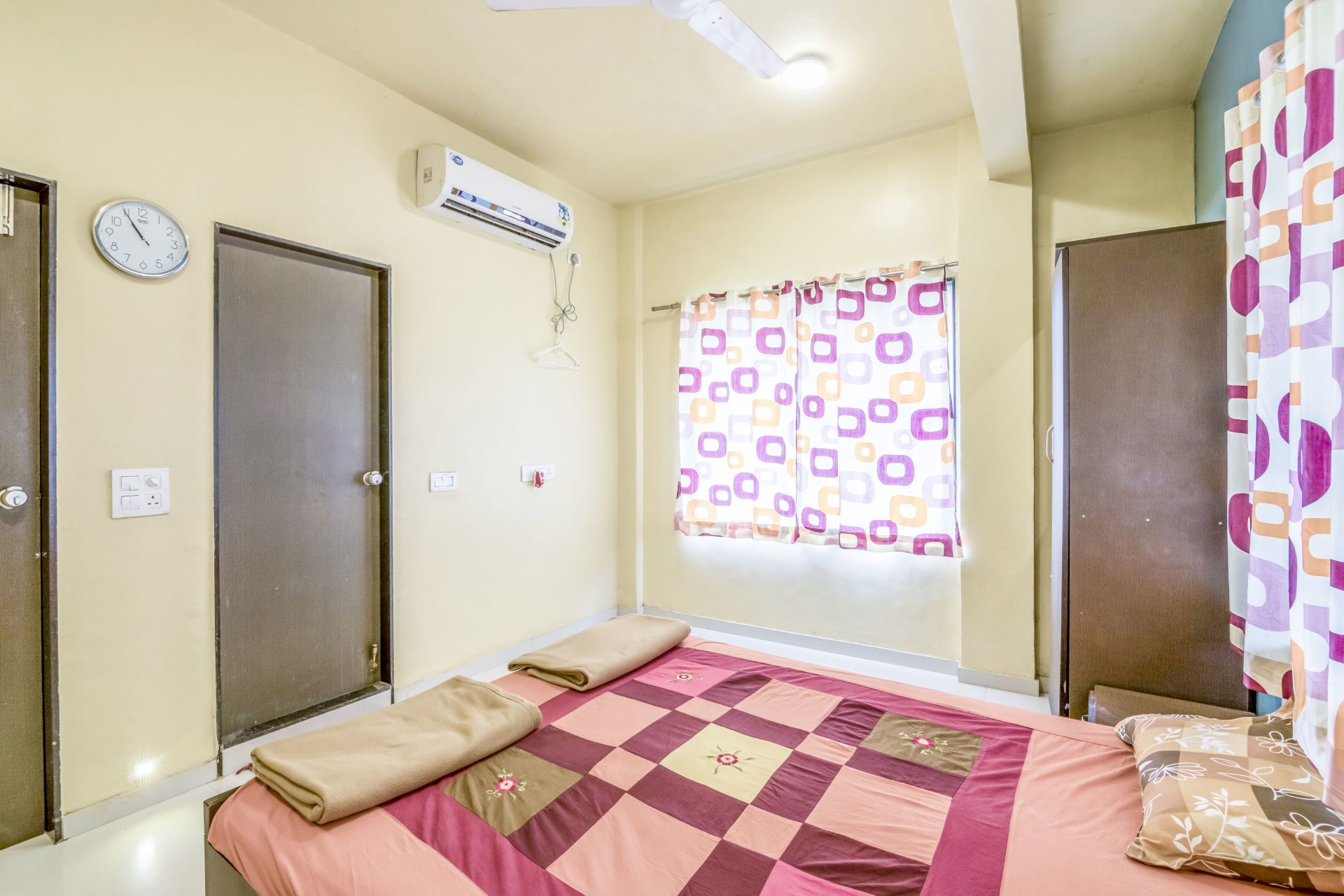 Une chambre à coucher avec un lit, deux portes, un climatiseur, une fenêtre avec des rideaux à motifs et une horloge au mur.