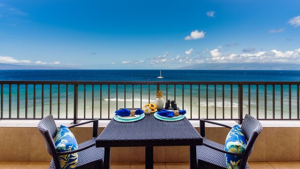 A balcony with a table set for two, overlooking a beach with a clear view of the ocean and a distant island.