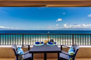 A balcony with a table set for two, overlooking a beach with a clear view of the ocean and a distant island. - Maui Kai 1005 1 Bedroom Condo by RedAwning (Lahaina)