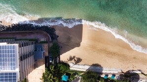 An aerial view of a beach with clear waters, a sandy shore, and a building with a rooftop terrace.