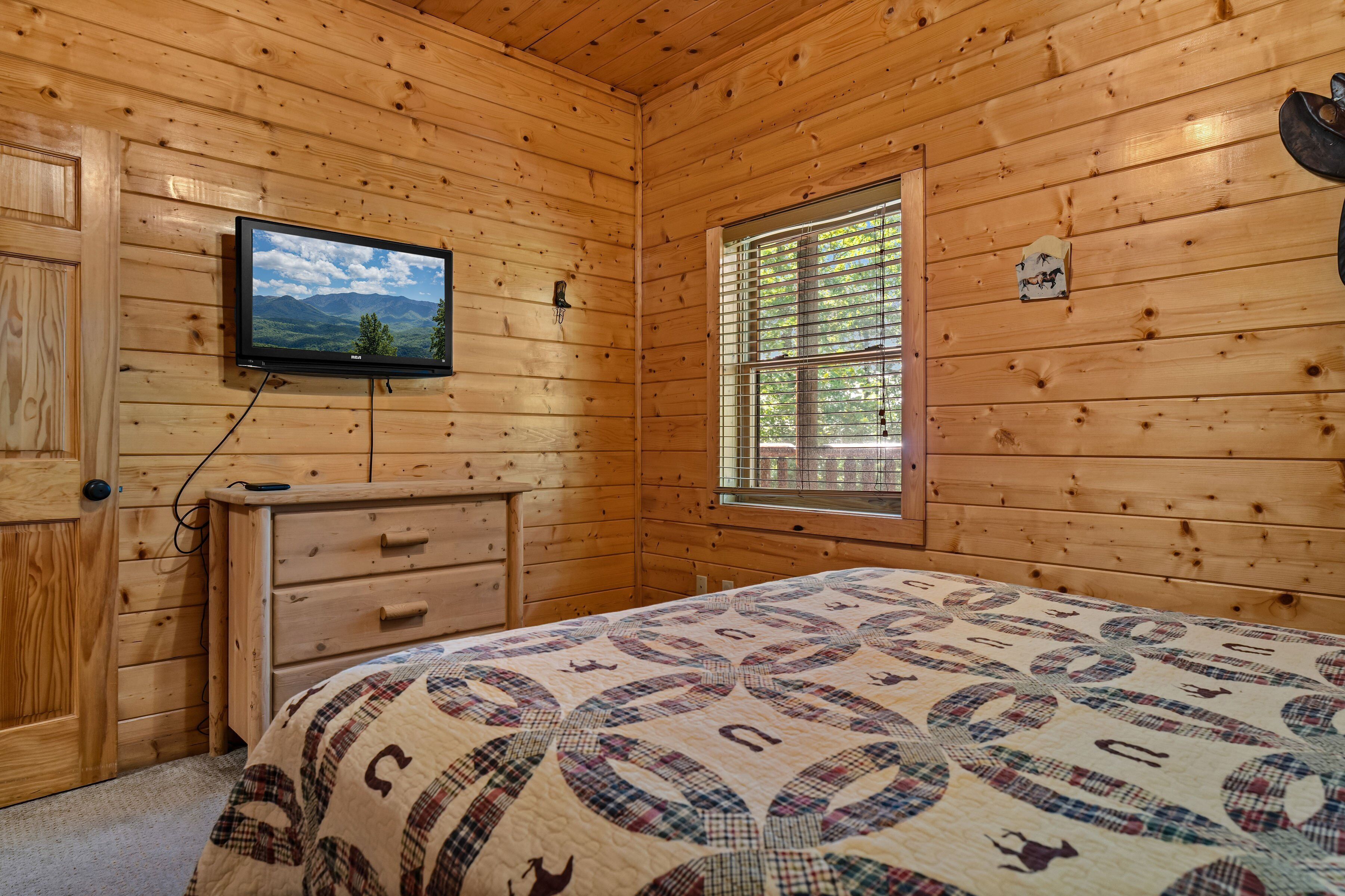 Una habitación de cabaña de madera con una cama, un aparador, un televisor y una ventana con persianas.