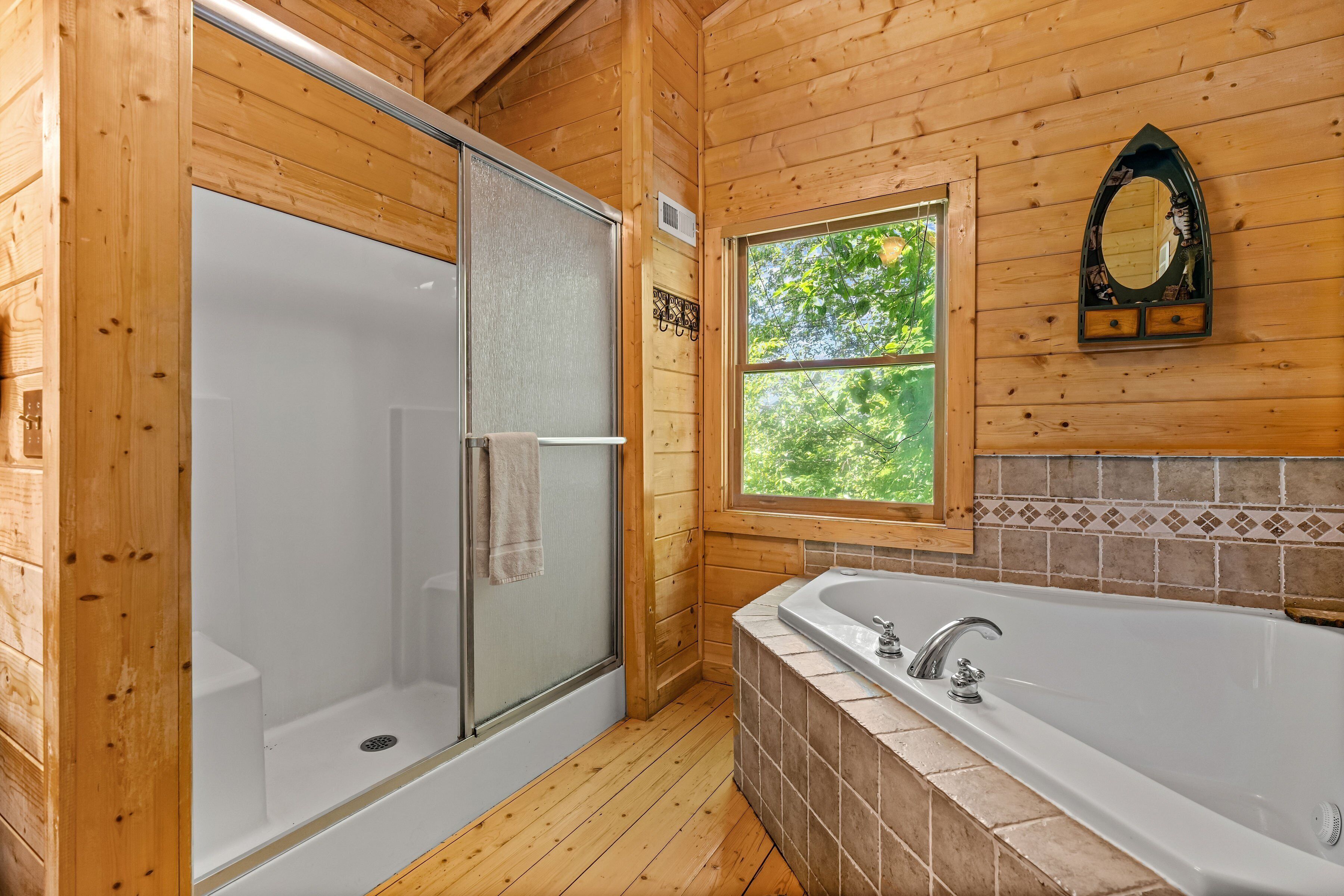 A wooden cabin bathroom with a glass-enclosed shower, a bathtub, and a window with a view of greenery.