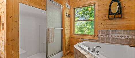 A wooden cabin bathroom with a glass-enclosed shower, a bathtub, and a window with a view of greenery.
