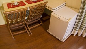Een kleine kamer met een houten tafel, twee stoelen, een kleine witte koelkast, een aan de muur bevestigde televisie en een airconditioner aan het plafond.