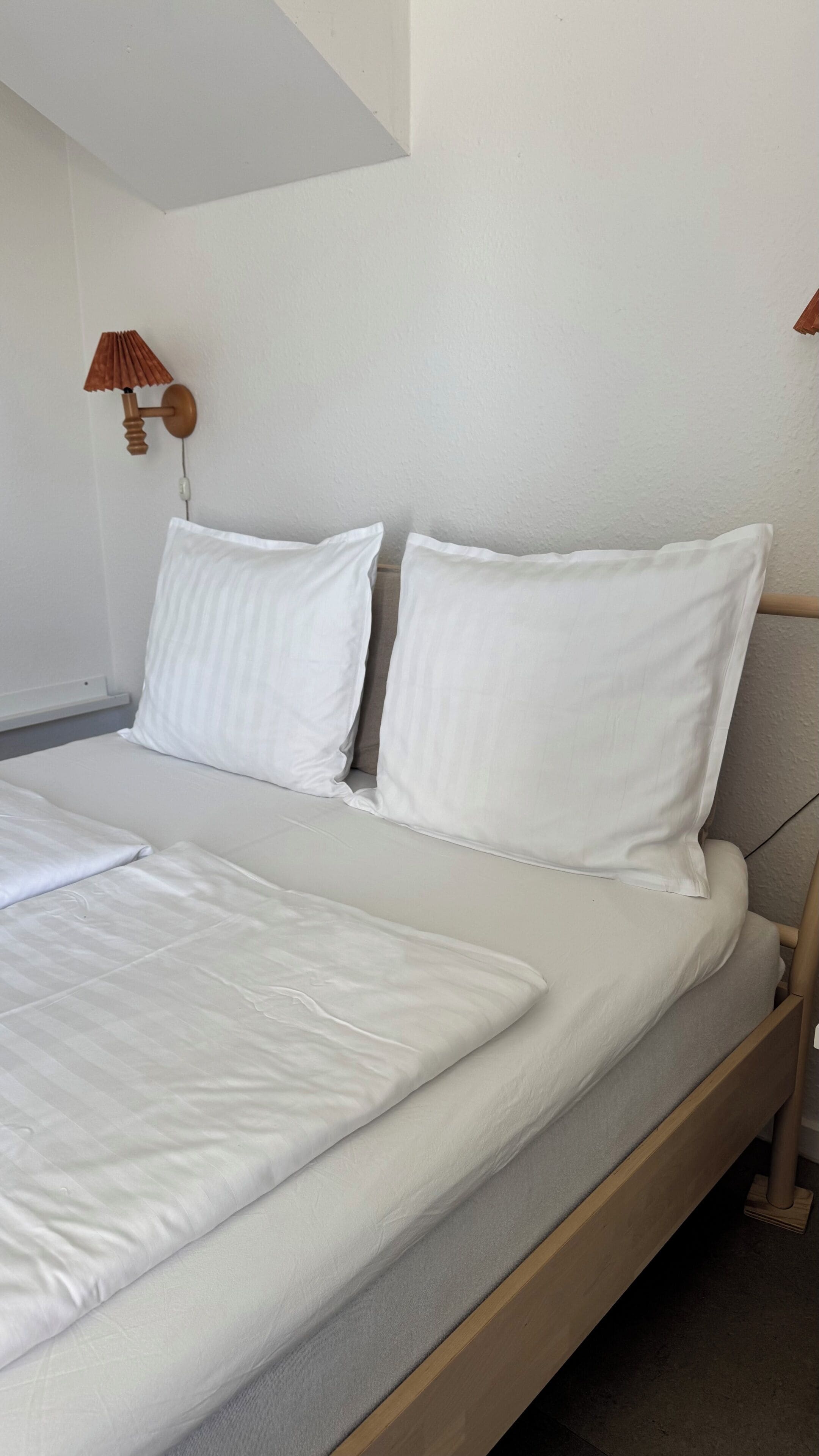 Ein ordentlich bezogenes Bett mit weißen Leinen, einem Holzkopfstück und einer Wandlampe.