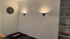 A room with a dartboard, two wall-mounted lights, a dart mat, and shelves with various items. - Hine Bakke Guest House (Rudkobing)