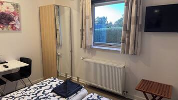 Un dormitorio con cama, escritorio, silla, radiador, ventana con cortinas y televisor en la pared.