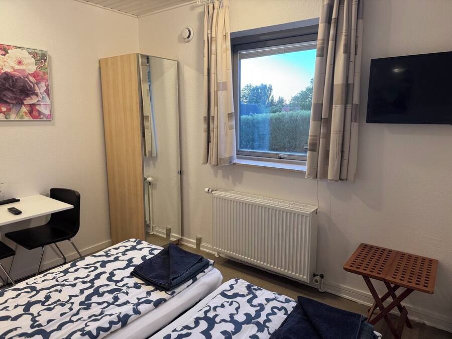 Quarto com cama, escrivaninha, cadeira, radiador, janela com cortinas e televisão na parede.
