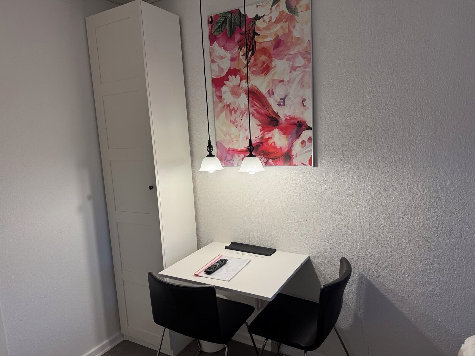 Et lille, hvidt skrivebord med to sorte stole, en hvidtavle og en fjernbetjening på bordet.