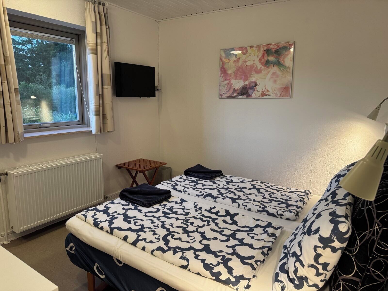 Makuuhuoneessa on sänky, pieni pöytä, televisio ja ikkuna, jossa on verhot.